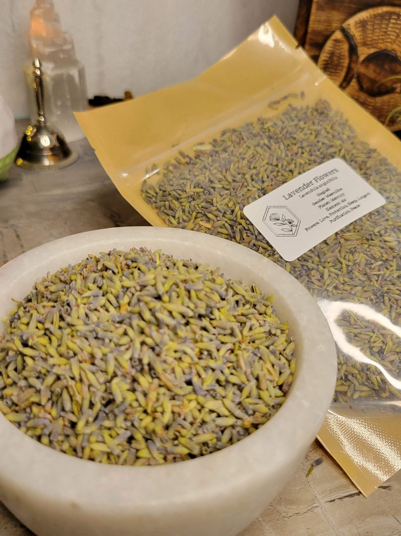 May include: A white ceramic bowl filled with dried lavender flowers. The flowers are a light purple and green colour. The bowl is sitting on a wooden surface. A clear plastic bag with a label that reads "Lavender Flowers" is in the background.