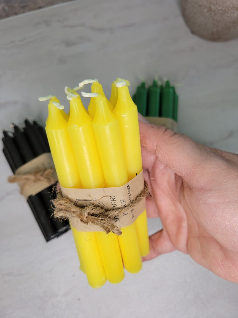 May include: A bundle of bright yellow taper candles secured with a brown paper band and tied with natural jute twine. The paper band features the text MY MAGIC PIECE and UNIQ HANDMADE. Additional bundles of black and green candles are visible nearby.
