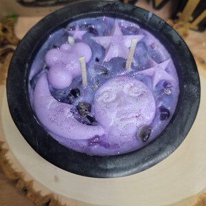 May include: A black cauldron-shaped candle filled with purple wax, featuring star, moon, and cloud-shaped wax accents. The candle is decorated with glitter and small crystals, and sits on a wooden slice.