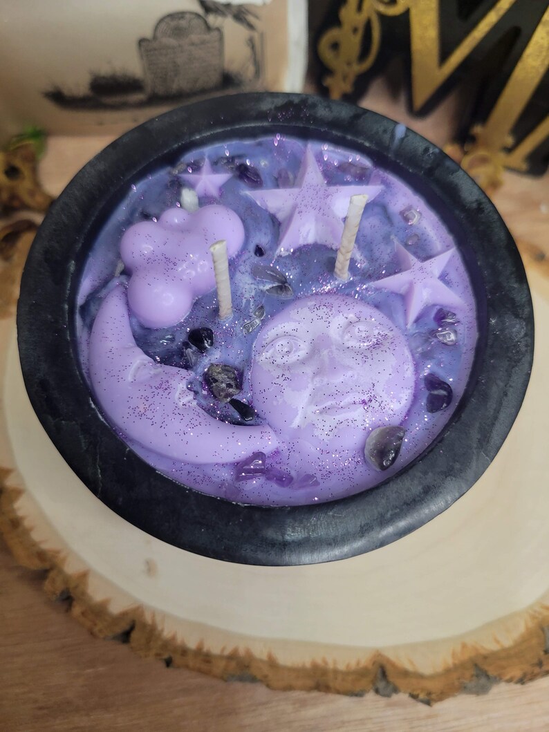 May include: A black cauldron-shaped candle filled with purple wax, featuring star, moon, and cloud-shaped wax accents. The candle is decorated with glitter and small crystals, and sits on a wooden slice.