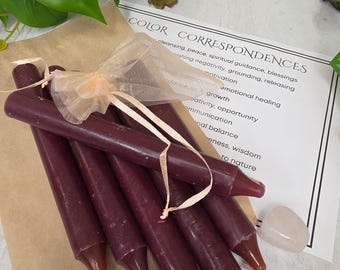 May include: A bundle of five dark burgundy taper candles tied together with a sheer peach-colored organza ribbon. The candles are placed on a brown paper sheet featuring printed text titled Color Correspondences, accompanied by a small smooth stone.