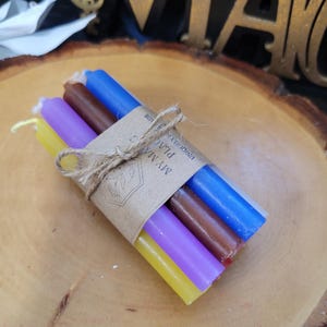 May include: A bundle of four thin taper candles in yellow, purple, brown, and blue, tied together with a piece of twine. A brown paper label is wrapped around the center, featuring a line drawing of a flower and the text MY MADE PLACE.