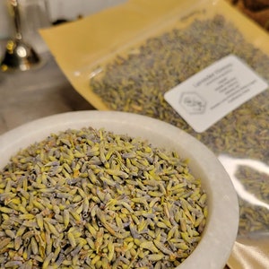 May include: A white ceramic bowl filled with dried lavender buds. The lavender buds are a mix of green and purple. The bowl is sitting on a wooden surface. The lavender buds are packaged in a clear plastic bag with a gold label that says "Lavender Flowers".
