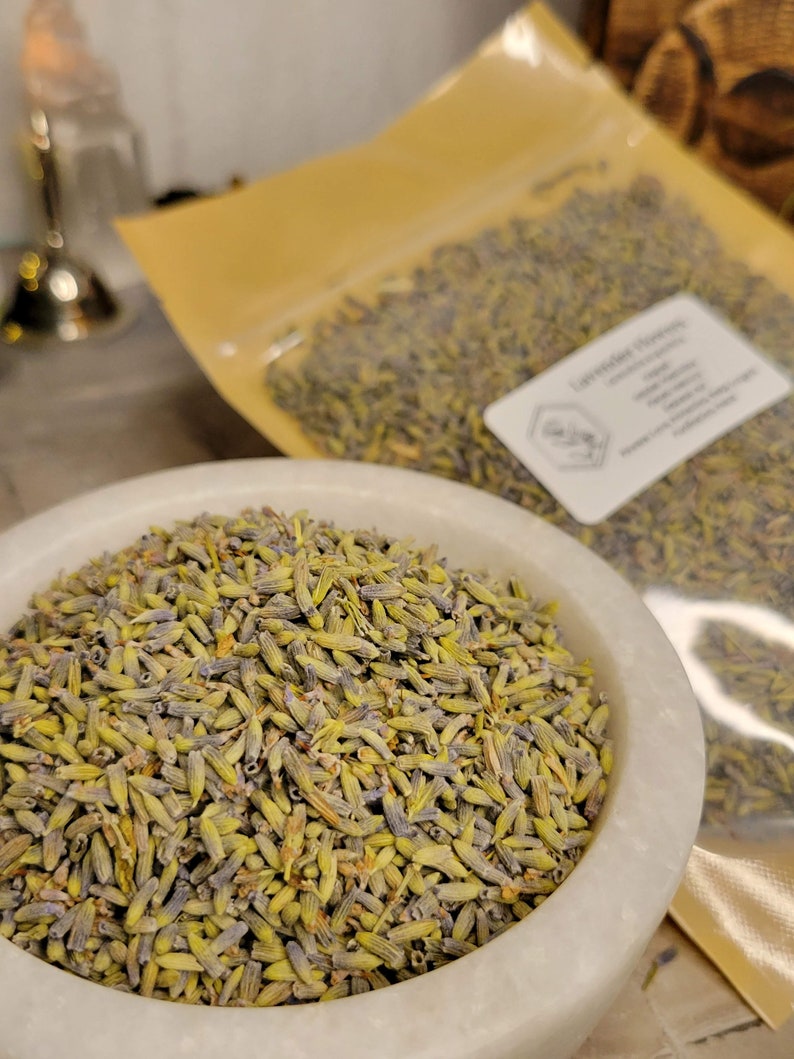 May include: A white ceramic bowl filled with dried lavender buds. The lavender buds are a mix of green and purple. The bowl is sitting on a wooden surface. The lavender buds are packaged in a clear plastic bag with a gold label that says "Lavender Flowers".