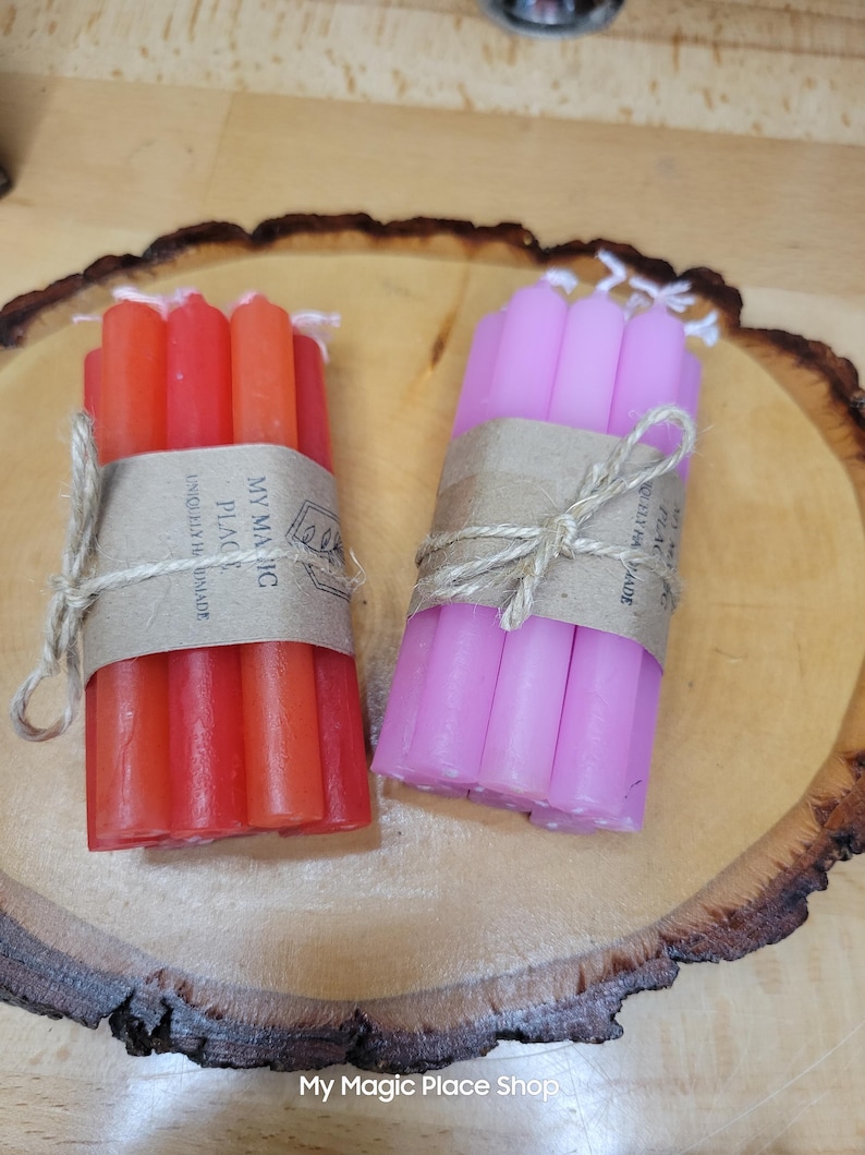 May include: Two bundles of thin taper candles, one set in shades of red and orange and the other in shades of pink and purple. Each bundle is wrapped in a brown paper band labeled My Magic Place Uniquely Handmade and tied with twine.