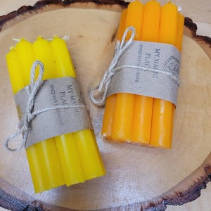 May include: Two bundles of thin taper candles, one set in bright yellow and one in deep orange. Each bundle is wrapped in a brown paper band labeled My Magic Place Uniquely Handmade and tied with a thin jute string, resting on a round wood slice.