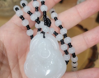 May include: White jade pendant of a smiling Buddha on a black and white beaded necklace. The pendant is carved with intricate details and has a smooth, polished finish.