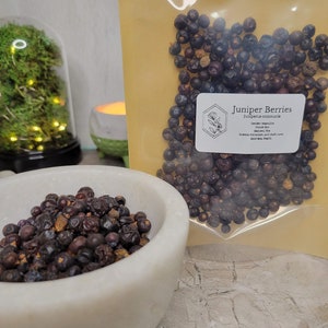 May include: A close-up of a white ceramic bowl filled with dried juniper berries. The berries are dark blue and black in color. A clear plastic bag filled with the same berries is in the background. The bag has a label that reads "Juniper Berries Juniperus communis Gender: Masculine Element: Fire Powers: Protection, Anti-theft, Love, Spiritual Health."