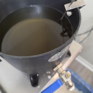 May include: A black metal melting pot with a blue-handled spigot and a silver candy thermometer clipped to the rim. The pot contains a translucent liquid and features a small circular logo on the side. The text My Magic Place Shop is visible.