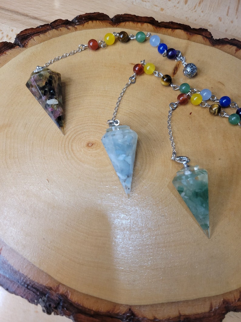 May include: Three crystal pendulums with silver chains hanging from a wooden surface. The pendulums are made of clear quartz, green aventurine, and amethyst.