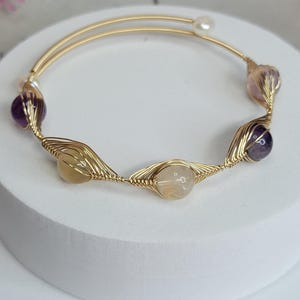May include: A gold-toned wire bracelet featuring a series of colorful gemstones. The bracelet has a delicate design with amethyst, rose quartz, and yellow gemstones. A small pearl is also incorporated into the design. The bracelet is displayed on a white surface.