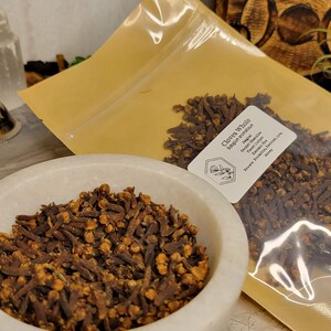 Puede incluir: Primer plano de una bolsa de clavos enteros, con una etiqueta que dice "Clavos enteros" y el nombre bot&aacute;nico "Syzygium aromaticum". Los clavos son marrones y tienen un ligero tono rojizo. La bolsa est&aacute; sobre una superficie blanca.