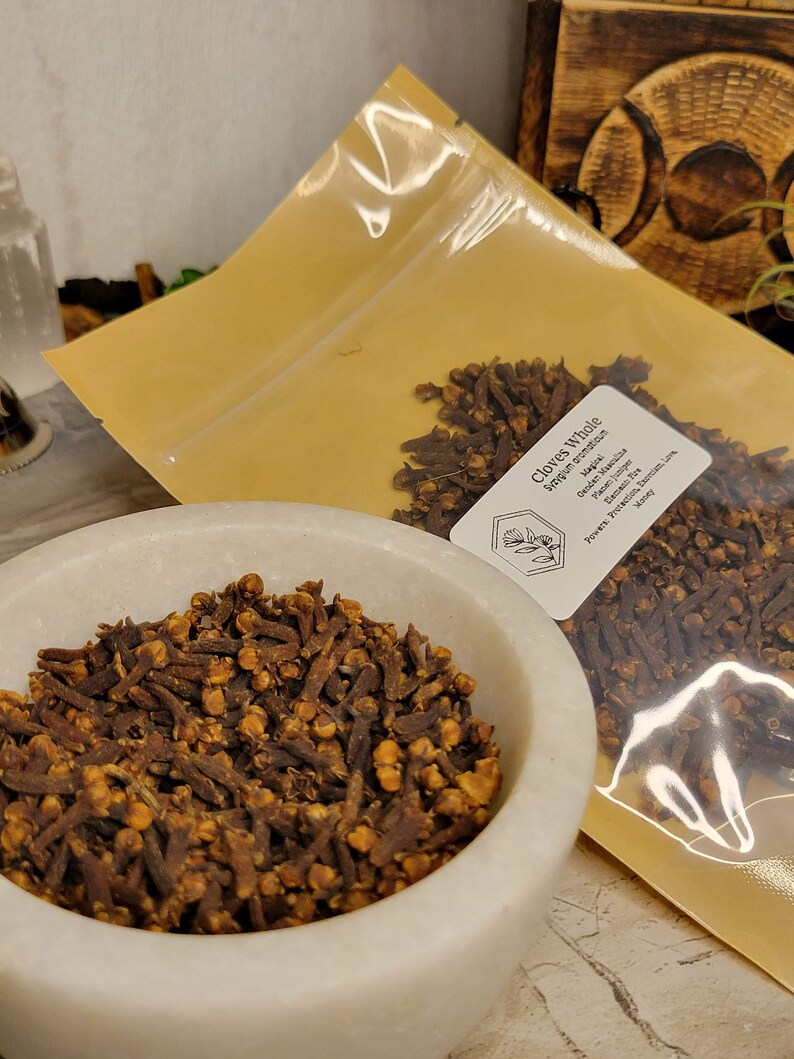 Puede incluir: Primer plano de una bolsa de clavos enteros, con una etiqueta que dice "Clavos enteros" y el nombre bot&aacute;nico "Syzygium aromaticum". Los clavos son marrones y tienen un ligero tono rojizo. La bolsa est&aacute; sobre una superficie blanca.
