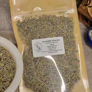 May include: A clear plastic bag filled with dried lavender flowers. The bag has a label that reads "Lavender Flowers" and includes information about the plant's magical properties, including its gender, planet, element, and powers.