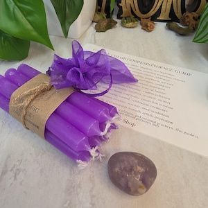 May include: A bundle of six slender purple taper candles wrapped in a brown paper band labeled My Magic Place, tied with a purple organza ribbon. Beside the candles is a smooth, polished purple stone and a printed Candle Color Correspondence Guide.