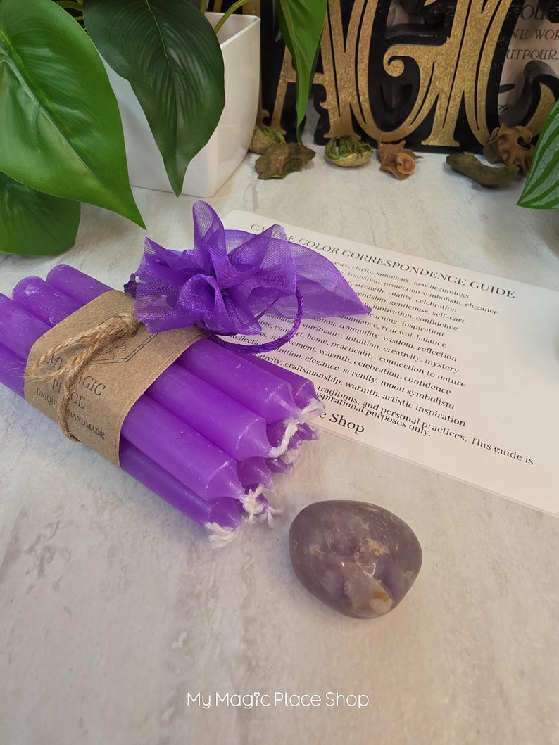 May include: A bundle of six slender purple taper candles wrapped in a brown paper band labeled My Magic Place, tied with a purple organza ribbon. Beside the candles is a smooth, polished purple stone and a printed Candle Color Correspondence Guide.