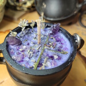 May include: A black cauldron-shaped candle with a lavender-colored wax, adorned with amethyst crystals, dried lavender, and glitter. The candle has a single wick and is set on a wooden surface. The cauldron has three small feet and a handle.