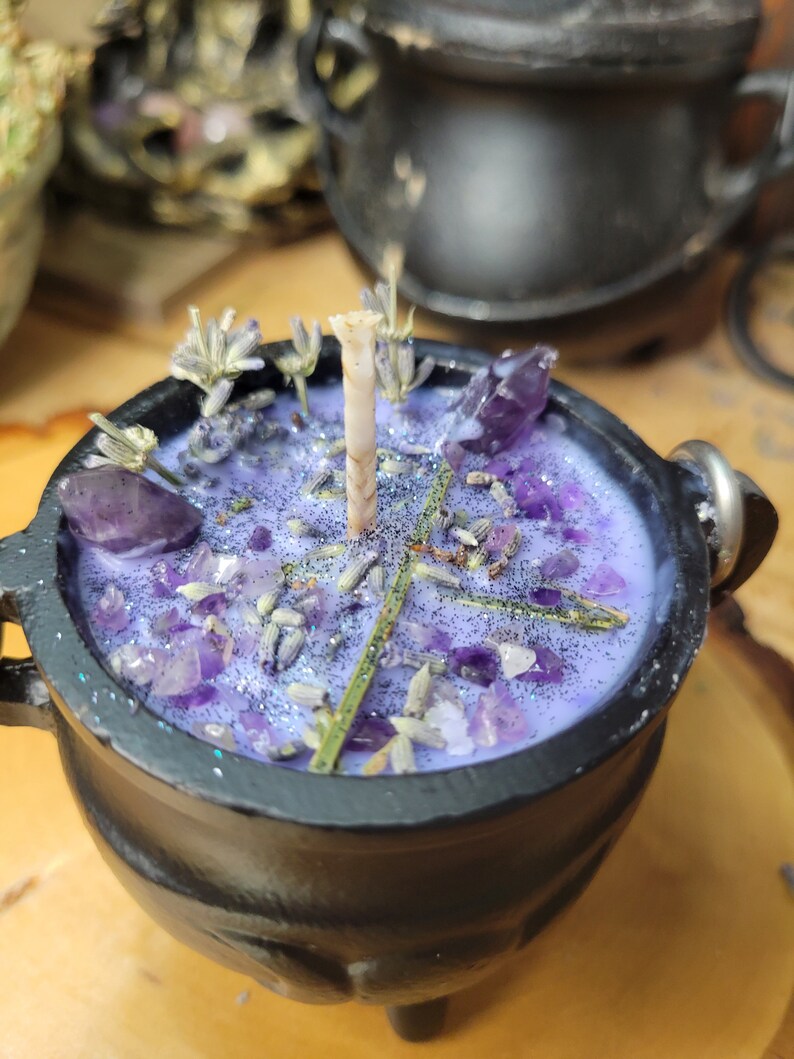 May include: A black cauldron-shaped candle with a lavender-colored wax, adorned with amethyst crystals, dried lavender, and glitter. The candle has a single wick and is set on a wooden surface. The cauldron has three small feet and a handle.