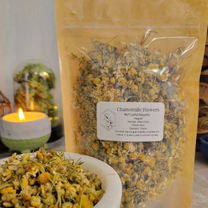 May include: A close-up of a bag of dried chamomile flowers. The bag is labeled "Chamomile Flowers Matricaria Recutita" and lists the magical properties of the herb, including its gender, planet, element, and powers. The flowers are a pale yellow color and have a delicate, floral scent.