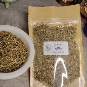 May include: A close-up of a bag of dried Agrimony herb. The bag is labeled with the name of the herb, its magical properties, and its planetary association. The label reads: "Agrimony, Agrimonia eupatoria, Magical, Gender: Masculine, Planet: Juniper, Element: Air, Powers: Protection, Sleep."