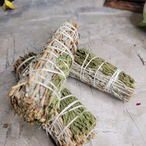 Pu&ograve; includere: Due mazzi di salvia e cedro legati con spago bianco. I mazzi sono impilati uno sopra l'altro e si trovano su una superficie grigia.