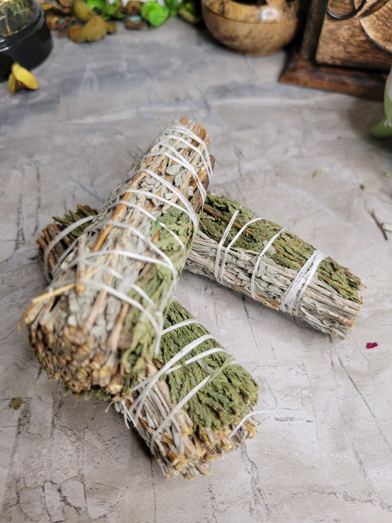 Pu&ograve; includere: Due mazzi di salvia e cedro legati con spago bianco. I mazzi sono impilati uno sopra l'altro e si trovano su una superficie grigia.