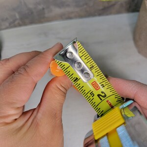 May include: A small, round, orange object being measured with a yellow metal tape measure. The tape measure shows the object has a diameter of approximately 0.5 inches. The tape measure features black markings and the text 25ft and Stanley.