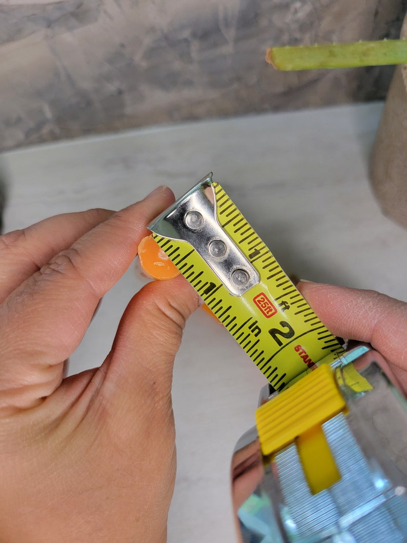 May include: A small, round, orange object being measured with a yellow metal tape measure. The tape measure shows the object has a diameter of approximately 0.5 inches. The tape measure features black markings and the text 25ft and Stanley.
