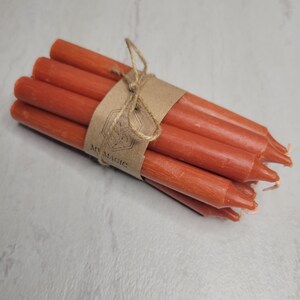 May include: A bundle of six thin, burnt-orange taper candles tied together with a brown paper band featuring a hexagonal logo and the text MY MAGIC. The candles have a smooth, matte wax finish and are arranged in a compact, cylindrical cluster.