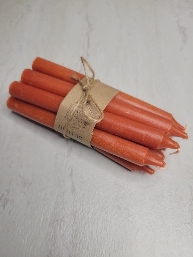 May include: A bundle of six thin, burnt-orange taper candles tied together with a brown paper band featuring a hexagonal logo and the text MY MAGIC. The candles have a smooth, matte wax finish and are arranged in a compact, cylindrical cluster.
