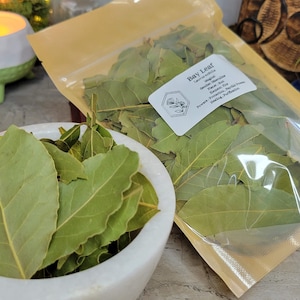 May include: A clear plastic bag filled with dried bay leaves. The bag is labeled "Bay Leaf" and lists the plant's magical properties, including protection, purification, and healing.
