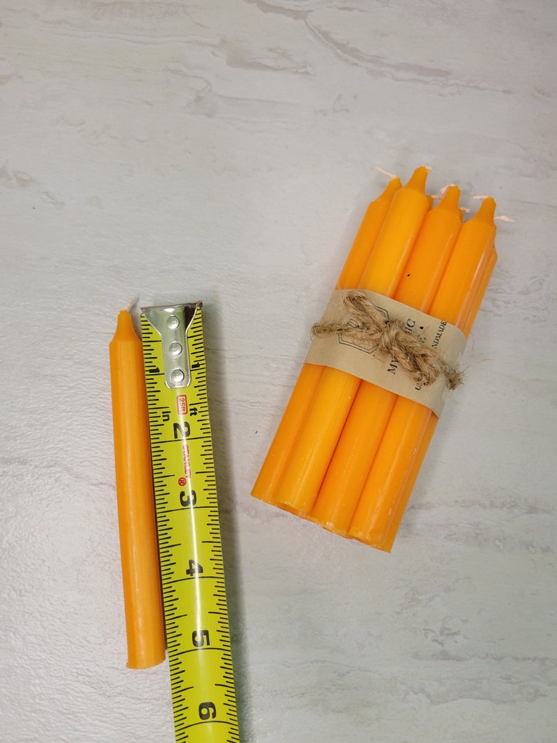 May include: A bundle of five smooth orange taper candles secured with a brown paper band and twine, positioned next to a yellow Stanley measuring tape showing a length of approximately six inches for each individual candle.