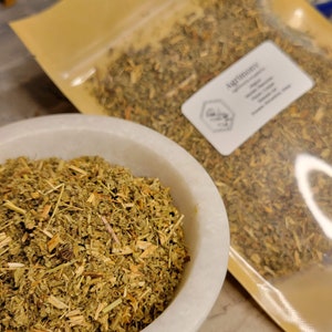 May include: A close-up of a white ceramic bowl filled with dried, light green and brown Agrimony herb. The herb is packaged in a clear plastic bag with a brown paper backing. The bag has a label that reads "Agrimony" and lists the herb's properties.