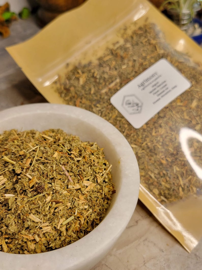 May include: A close-up of a white ceramic bowl filled with dried, light green and brown Agrimony herb. The herb is packaged in a clear plastic bag with a brown paper backing. The bag has a label that reads "Agrimony" and lists the herb's properties.