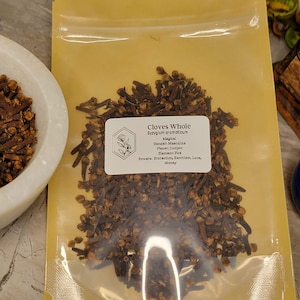 Puede incluir: Un primer plano de una peque&ntilde;a bolsa de pl&aacute;stico transparente llena de clavos de olor enteros. La bolsa est&aacute; etiquetada como "Clavos de olor enteros Syzygium aromaticum" y enumera las propiedades m&aacute;gicas de los clavos de olor, incluida la protecci&oacute;n, el exorcismo, el amor y el dinero.