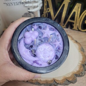 May include: A black cauldron-shaped candle with a lavender wax design featuring a smiling moon, stars, and a cloud. The candle is decorated with purple crystals and glitter. The candle is on a wooden surface.