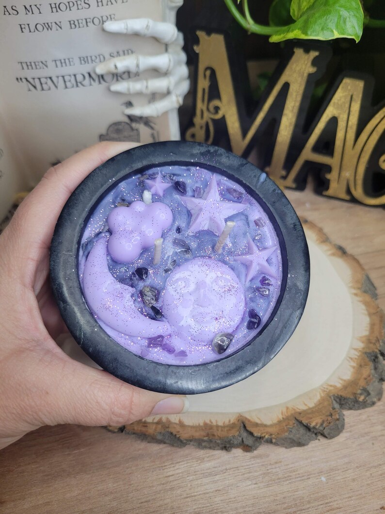 May include: A black cauldron-shaped candle with a lavender wax design featuring a smiling moon, stars, and a cloud. The candle is decorated with purple crystals and glitter. The candle is on a wooden surface.
