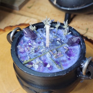 May include: A black cauldron-shaped candle with a lavender-colored wax. The candle is decorated with amethyst crystals, dried lavender sprigs, and glitter. A white candle wick is centered in the wax. The cauldron has three small legs and two metal handles.