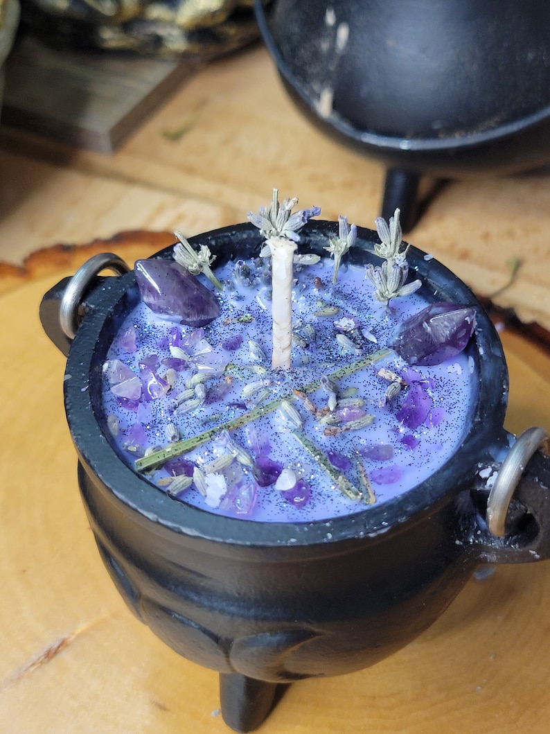 May include: A black cauldron-shaped candle with a lavender-colored wax. The candle is decorated with amethyst crystals, dried lavender sprigs, and glitter. A white candle wick is centered in the wax. The cauldron has three small legs and two metal handles.