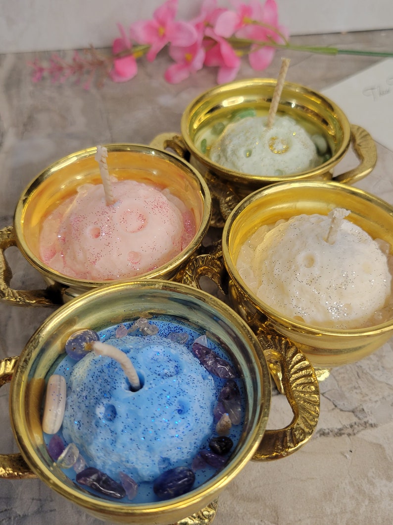 May include: Four decorative candles in gold-colored teacups. Each candle is a different color: pink, green, white, and blue. The blue candle is surrounded by purple and clear stones. The candles have wicks and glitter.