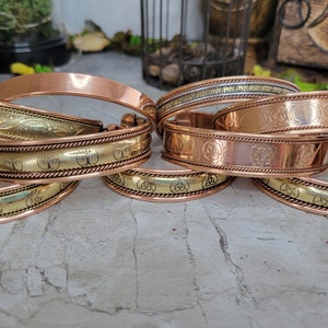 May include: A set of six copper bracelets with various engraved designs, including a tree of life, a triple moon, and a pentagram. The bracelets are stacked on top of each other, showcasing the different designs.