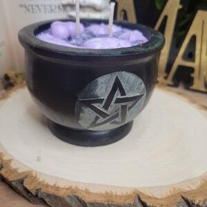 May include: A black cauldron-shaped candle with a pentagram design. The candle has a purple wax interior with two wicks. The cauldron sits on a wooden slice. The candle is a decorative item.