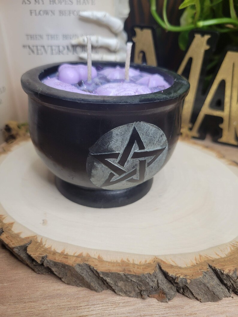 May include: A black cauldron-shaped candle with a pentagram design. The candle has a purple wax interior with two wicks. The cauldron sits on a wooden slice. The candle is a decorative item.