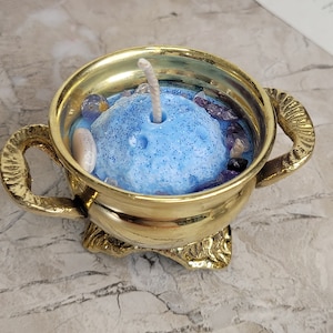 May include: A gold-colored candle holder with a blue candle. The candle is decorated with small stones and glitter. The candle holder has two handles and a decorative base. The candle is centered in the holder.