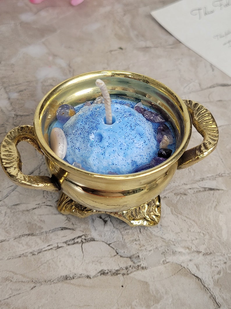 May include: A gold-colored candle holder with a blue candle. The candle is decorated with small stones and glitter. The candle holder has two handles and a decorative base. The candle is centered in the holder.