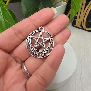 May include: A circular silver-toned metal pendant featuring a central five-pointed star enclosed within a crescent moon shape, with intricate Celtic knotwork detailing along the bottom curve and a small loop at the top for attachment.