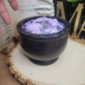 May include: A black cauldron-shaped candle filled with lavender wax, crystals, and decorative elements. The candle sits on a wooden slice. The text "NEVERMORE" is visible in the background.