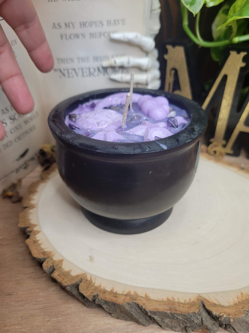 May include: A black cauldron-shaped candle filled with lavender wax, crystals, and decorative elements. The candle sits on a wooden slice. The text "NEVERMORE" is visible in the background.