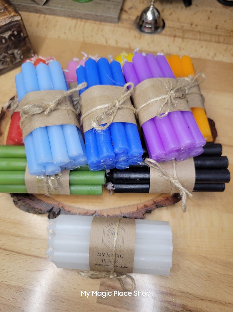 May include: A collection of multi-colored taper candles bundled with brown paper wraps and twine. The set includes light blue, dark blue, purple, orange, red, green, black, and white candles. A label on the white bundle reads My Magic Place.