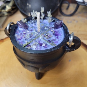 May include: A black cauldron-shaped candle with a purple wax interior. The candle is decorated with amethyst crystals, dried lavender, and a central wick. The cauldron has three small legs and two metal handles.